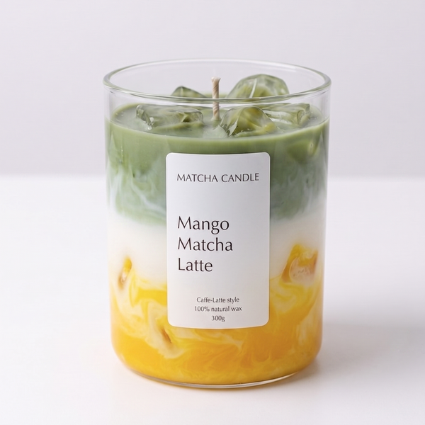 A glass jar candle with a white label reading 'MATCHA CANDLE' and 'Mango Matcha Latte'. The candle wax is layered with green matcha on top, white in the middle, and yellow-orange mango pieces on the bottom, with clear ice cubes floating in the wax.